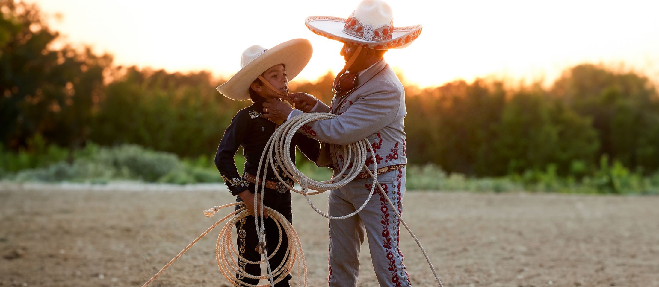 Un hombre y un niño vestidos con el traje tradicional Charro, sosteniendo cuerdas y parados uno al lado del otro.